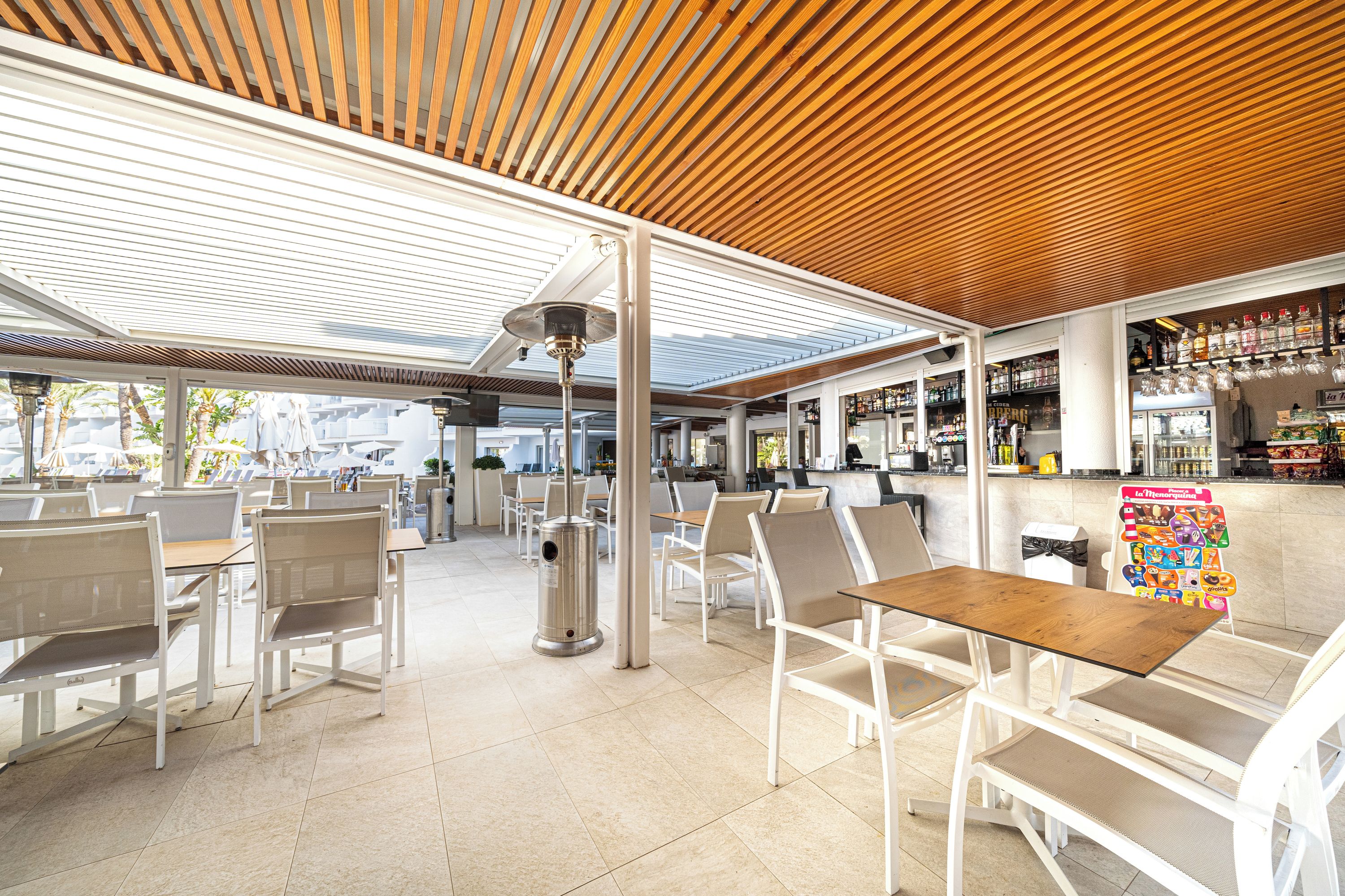 Un café en plein air avec des meubles blancs, un plafond en bois et un bar avec des étagères remplies de bouteilles.