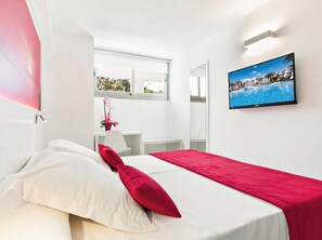 Een moderne hotelkamer met een bed, een rode plaid, witte kussens, een tafeltje met een bloevas, een spiegel en een televisie waarop een zwembad te zien is.