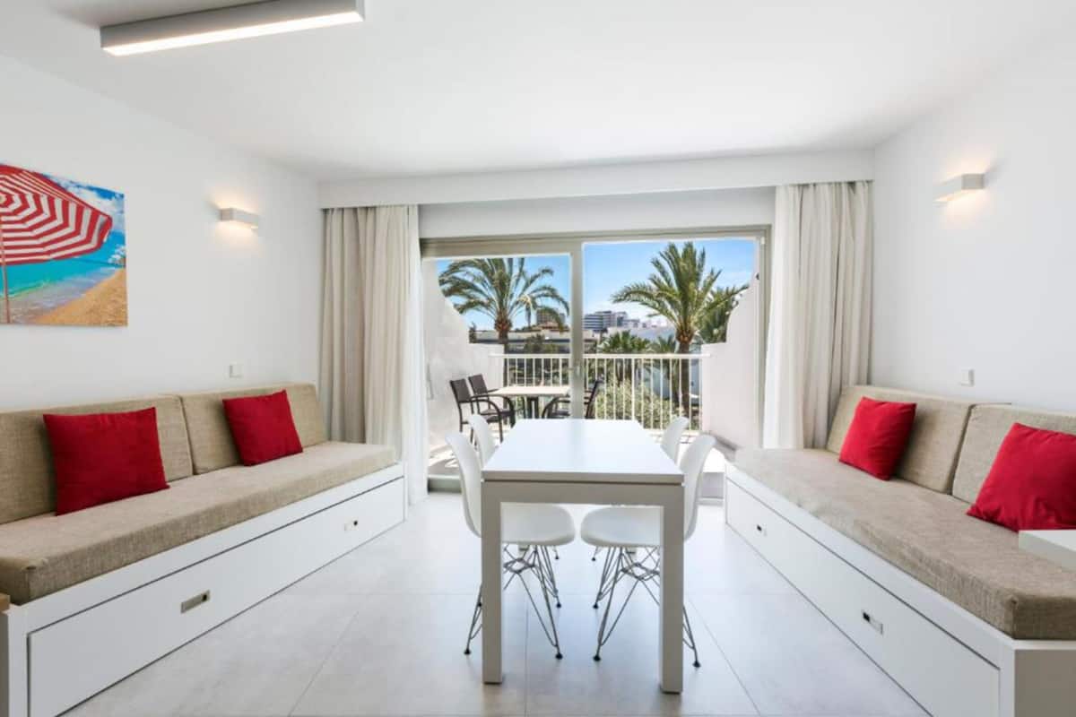 Une chambre d’hôtel moderne dotée d’un balcon, d’une table à manger et de deux canapés avec des coussins rouges.