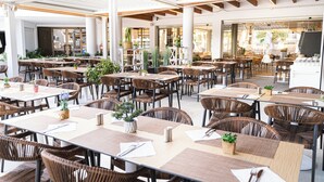 Een ruime eethoek buiten met meerdere tafels en rieten stoelen, omringd door grote ramen en potplanten.