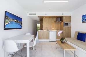 Een moderne keuken met een houten eiland, een eethoek met witte stoelen, een bank met een blauw kussen en een televisie waarop een zwembad te zien is.