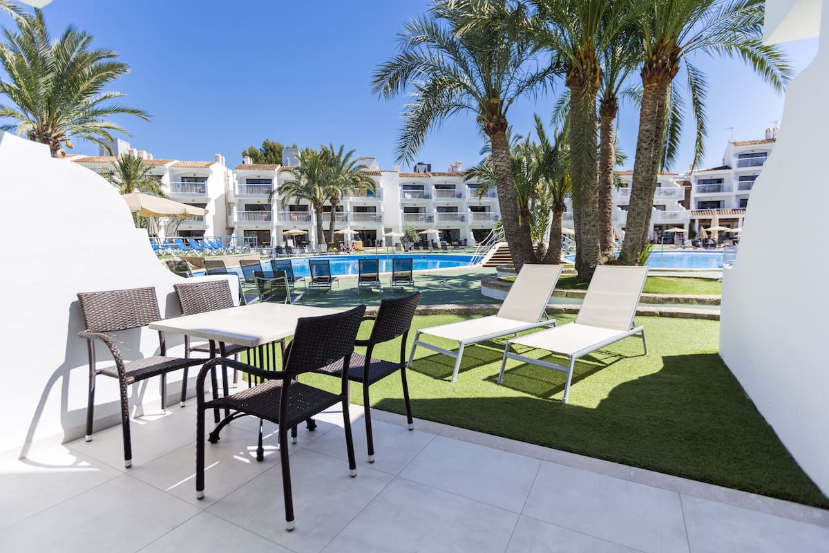 Un espace aménagé au bord de la piscine, avec des chaises longues et une table, entouré de palmiers et d’un bâtiment doté de balcons en arrière-plan.