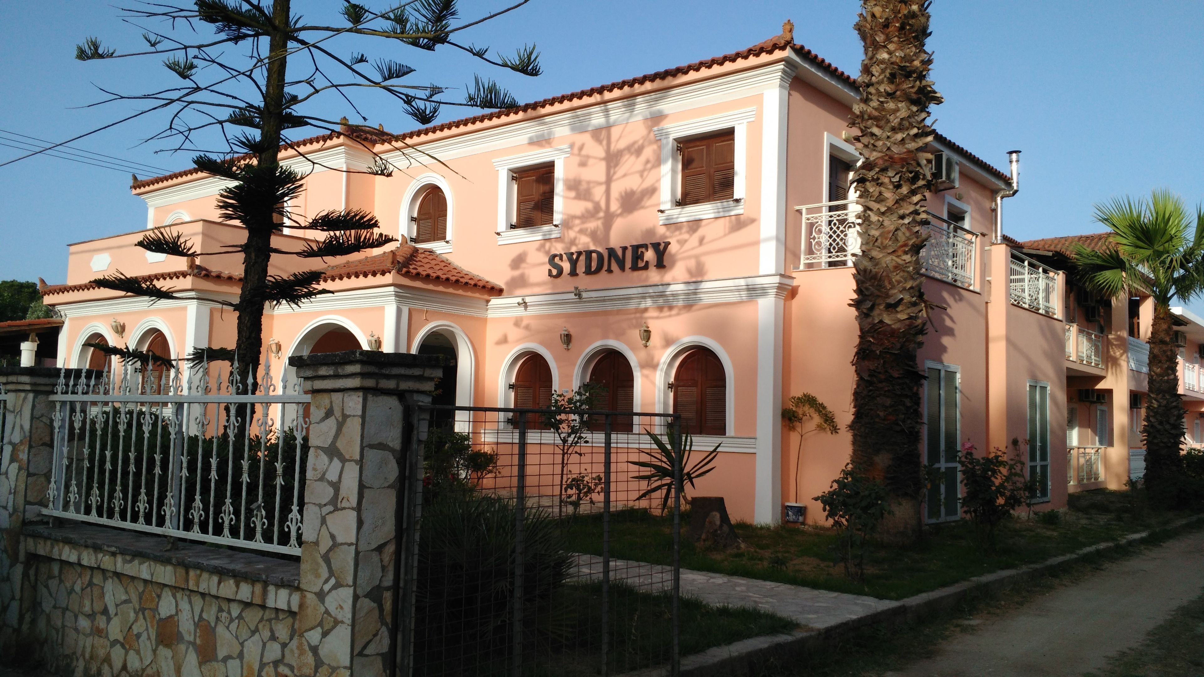 Un edificio con el nombre 'SYDNEY' en la fachada, que cuenta con un muro de piedra, una cerca de metal y una palmera.