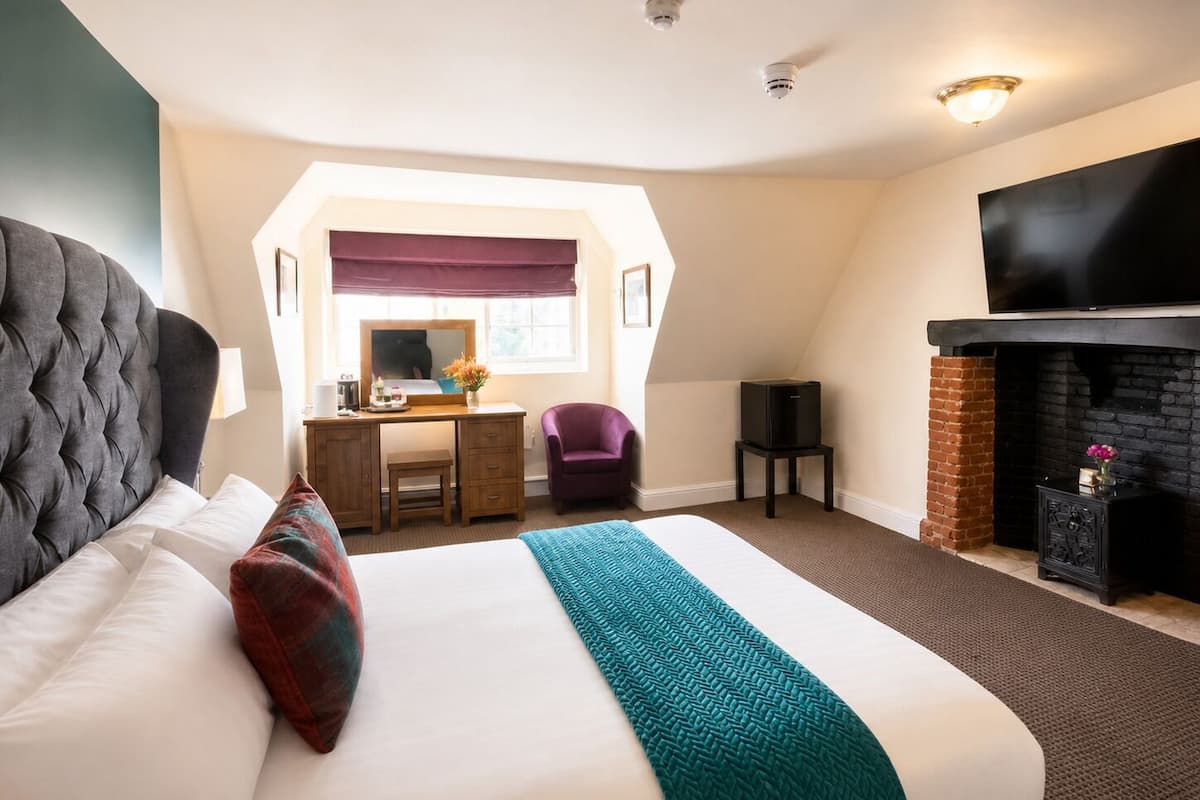 A hotel room with a large bed, a desk with a chair, a television, and a fireplace.