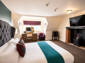 A hotel room with a large bed, a desk with a chair, a television, and a fireplace. - The Scole Hotel (Diss)