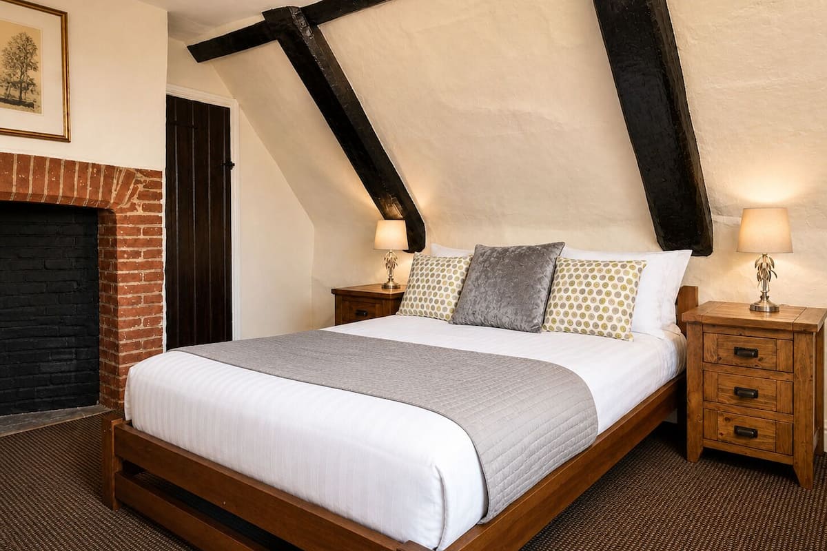 A bedroom with a sloped ceiling, a fireplace, a bed with a white duvet, patterned pillows, and two bedside tables with lamps.