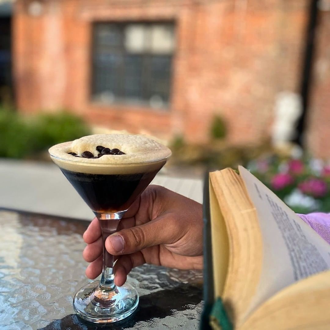 Une personne tenant un verre à martini contenant un liquide foncé surmonté d’une mousse, à côté d’un livre ouvert posé sur une table.