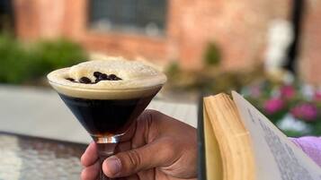 Une personne tenant un verre à martini contenant un liquide foncé surmonté d’une mousse, à côté d’un livre ouvert posé sur une table.
