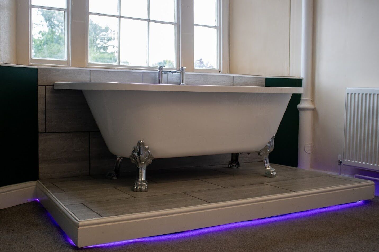 Une baignoire sur pieds dans une salle de bain avec une fenêtre au-dessus, un radiateur au mur et une bande lumineuse violette sur le sol.