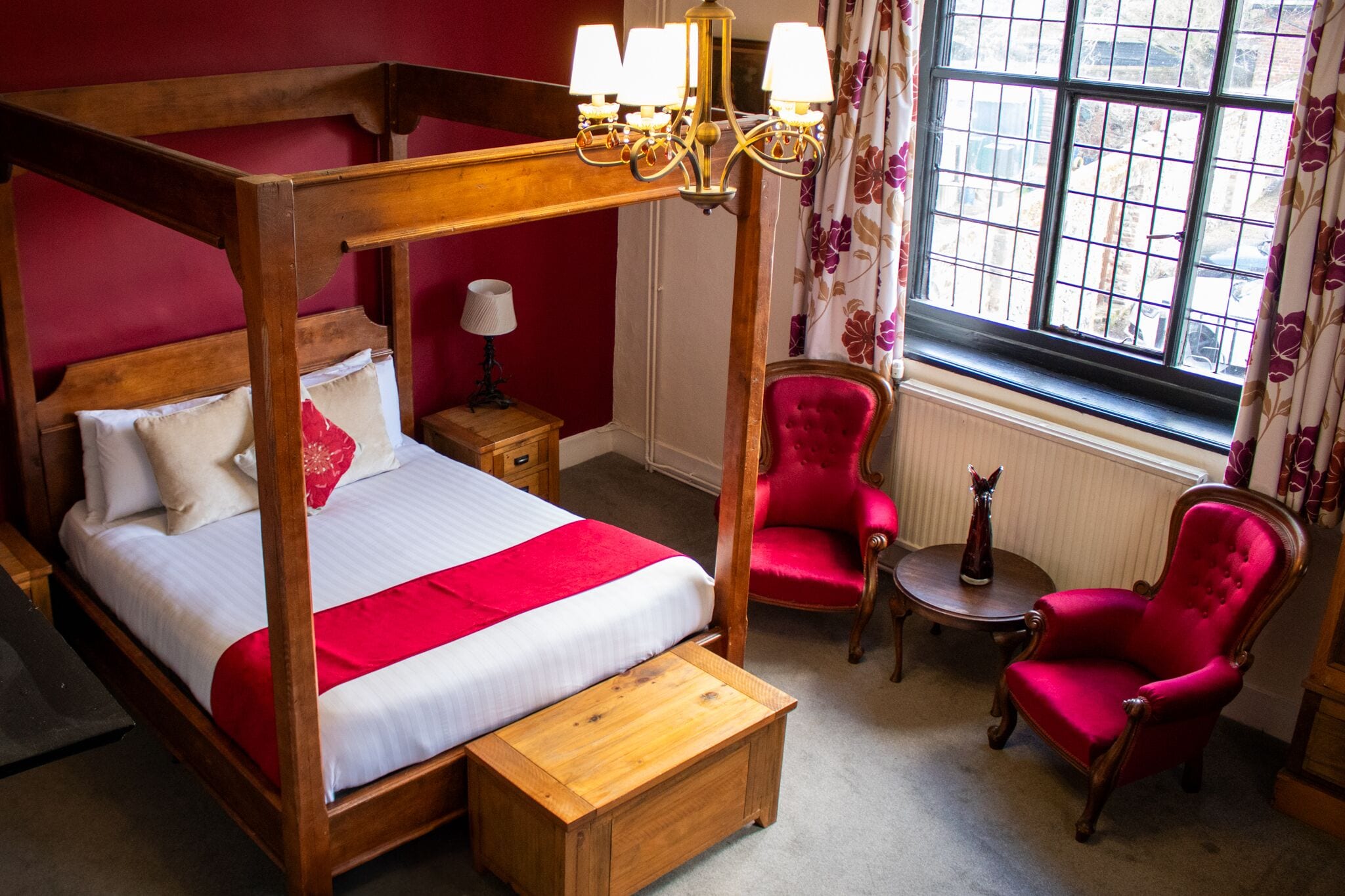 A four-poster bed with a wooden frame and white bedding, a red chair, a small round table, and a window with floral curtains.