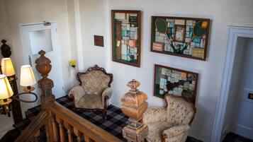 A staircase with a checkered floor, two upholstered chairs, a wooden table with a lamp, and framed pictures on the wall.