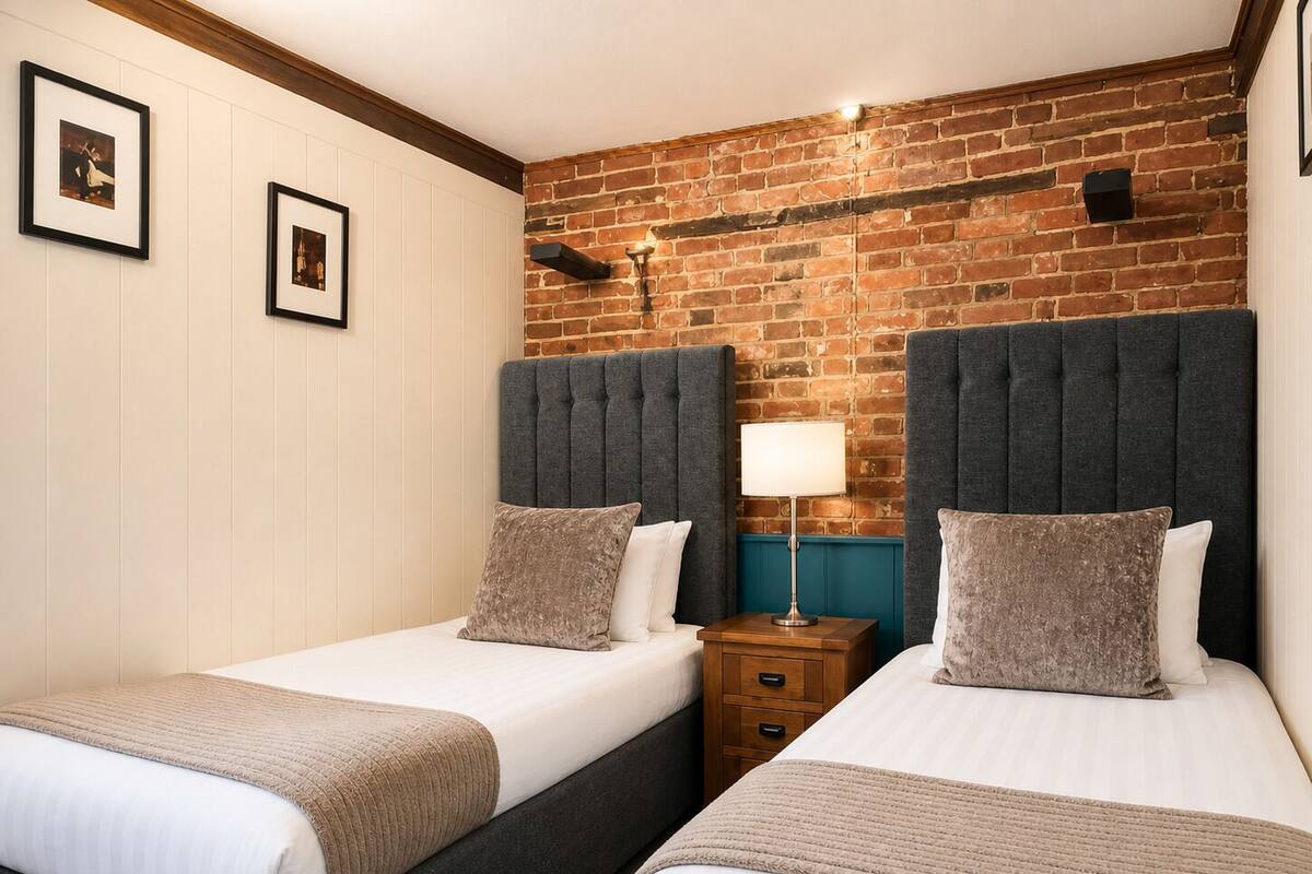 A hotel room with two beds, a brick wall, a wooden ceiling, and a nightstand with a lamp.