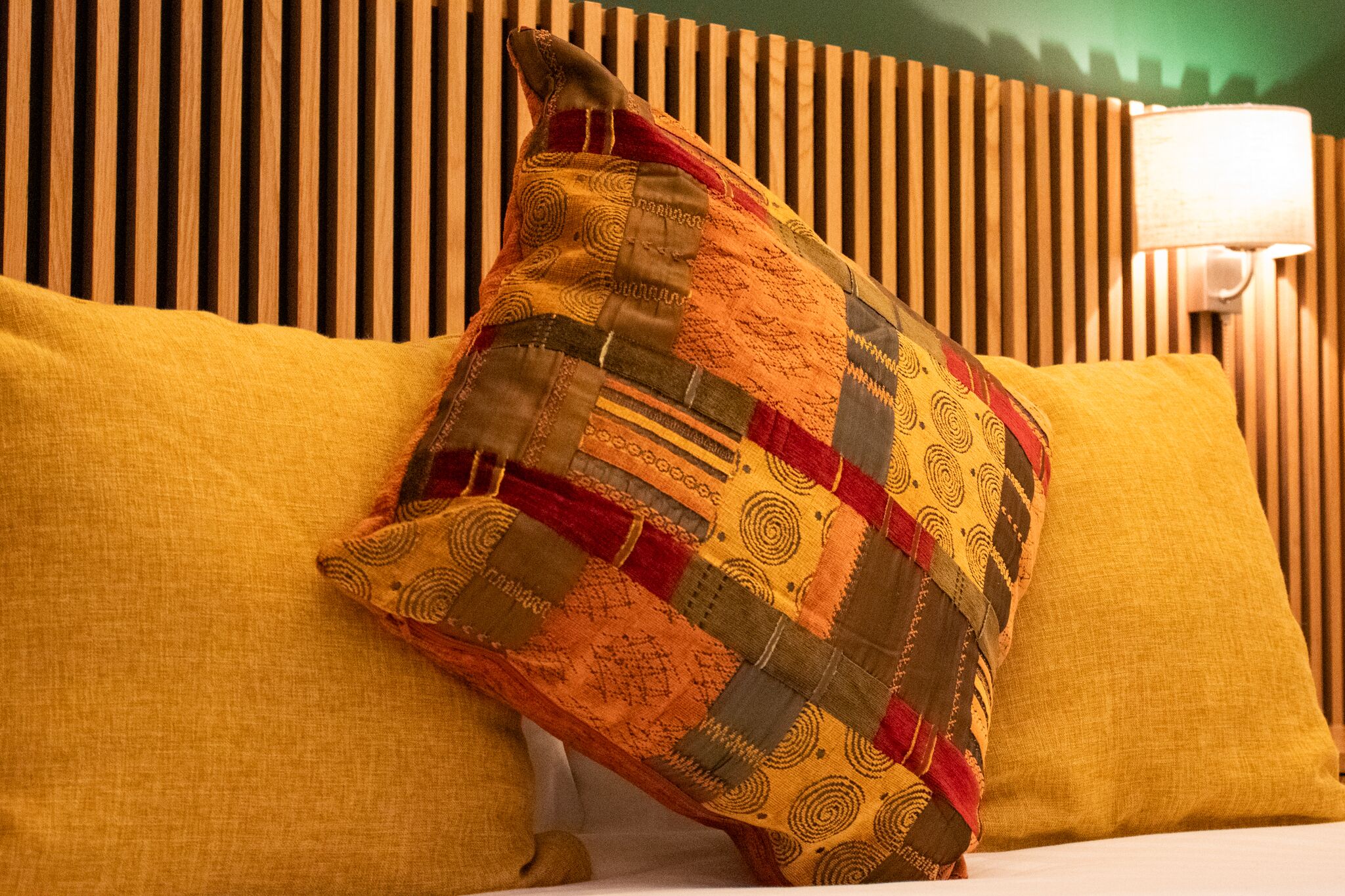Una cama con un almohadón amarillo y otro almohadón estampado, frente a un cabecero de madera.