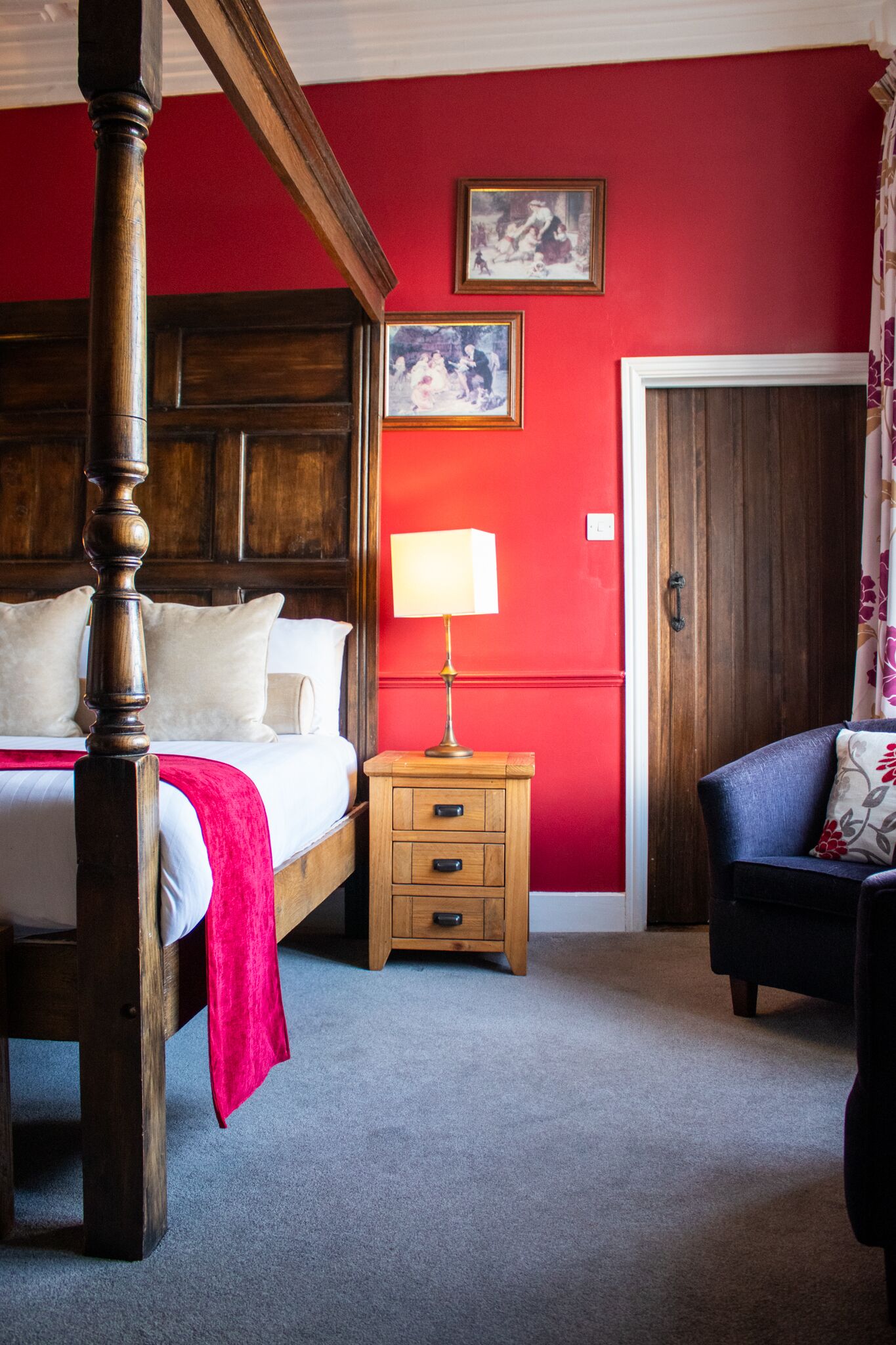 A bedroom with a four-poster bed, a nightstand with a lamp, a chair, and two framed pictures on the wall.