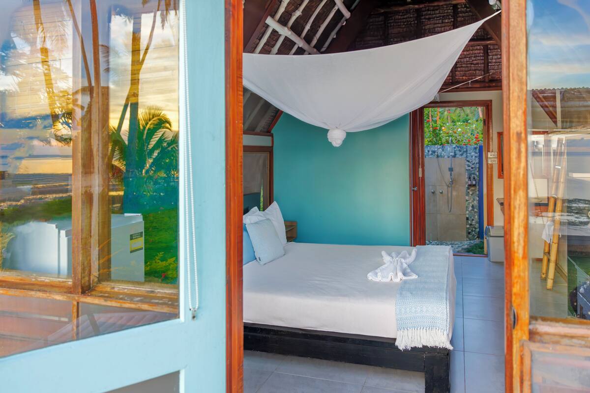 A room with a bed, a window with a view of palm trees, and a door leading outside.