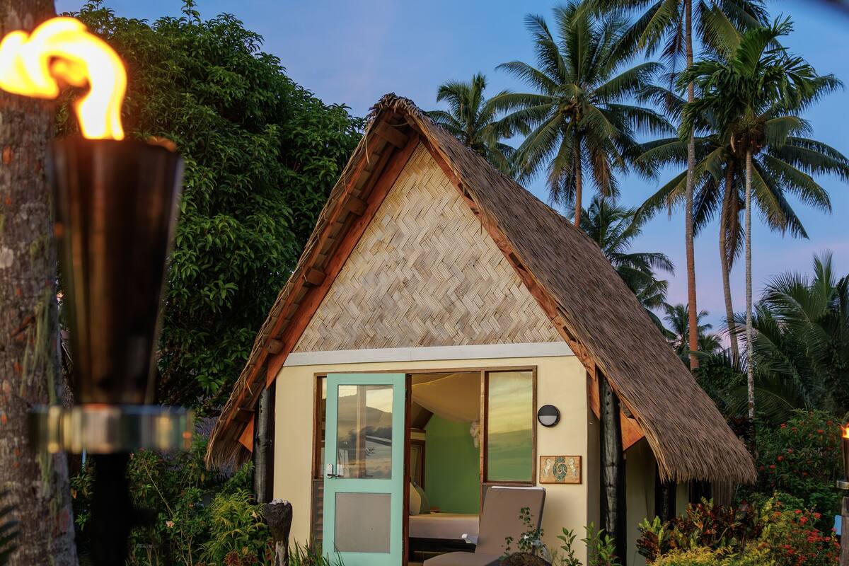 A thatched-roof bungalow with a door and a small porch, surrounded by tropical plants and palm trees.