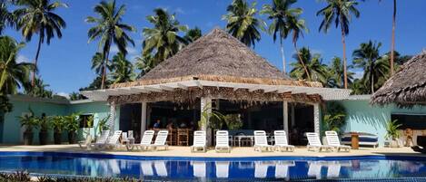 A poolside area with lounge chairs, a thatched-roof pavilion, and palm trees.