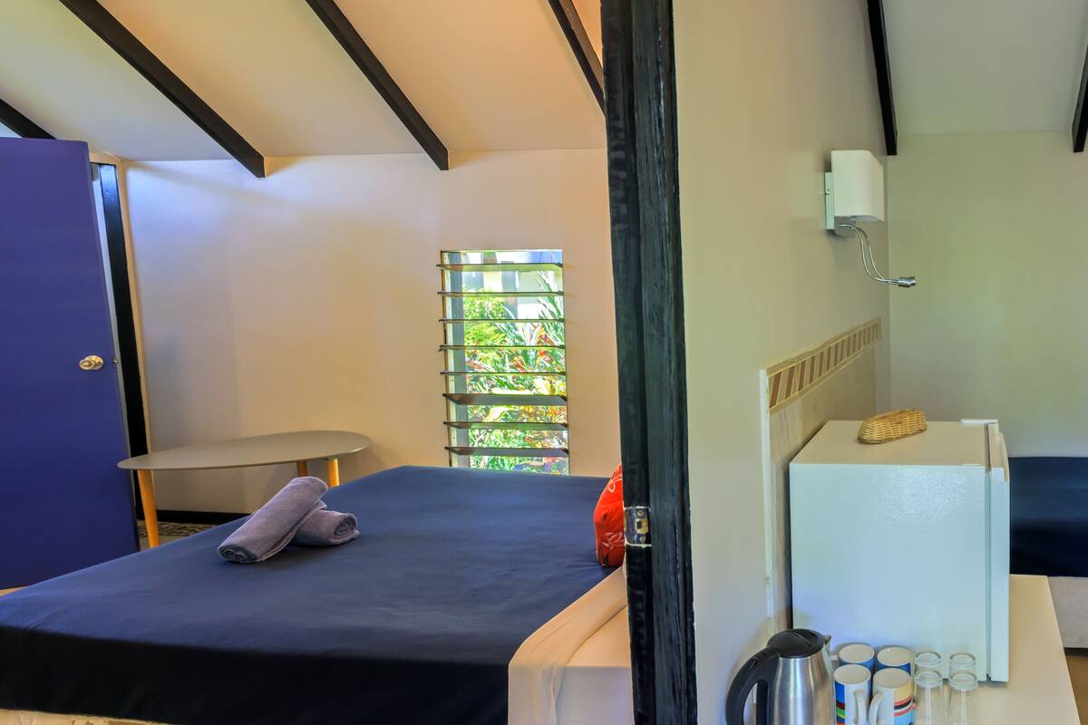 A room with two beds, a small table, a refrigerator, and a window with a view of greenery.