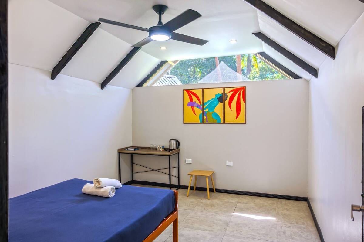 A small, well-lit room with a bed, a desk, a chair, and a painting on the wall.