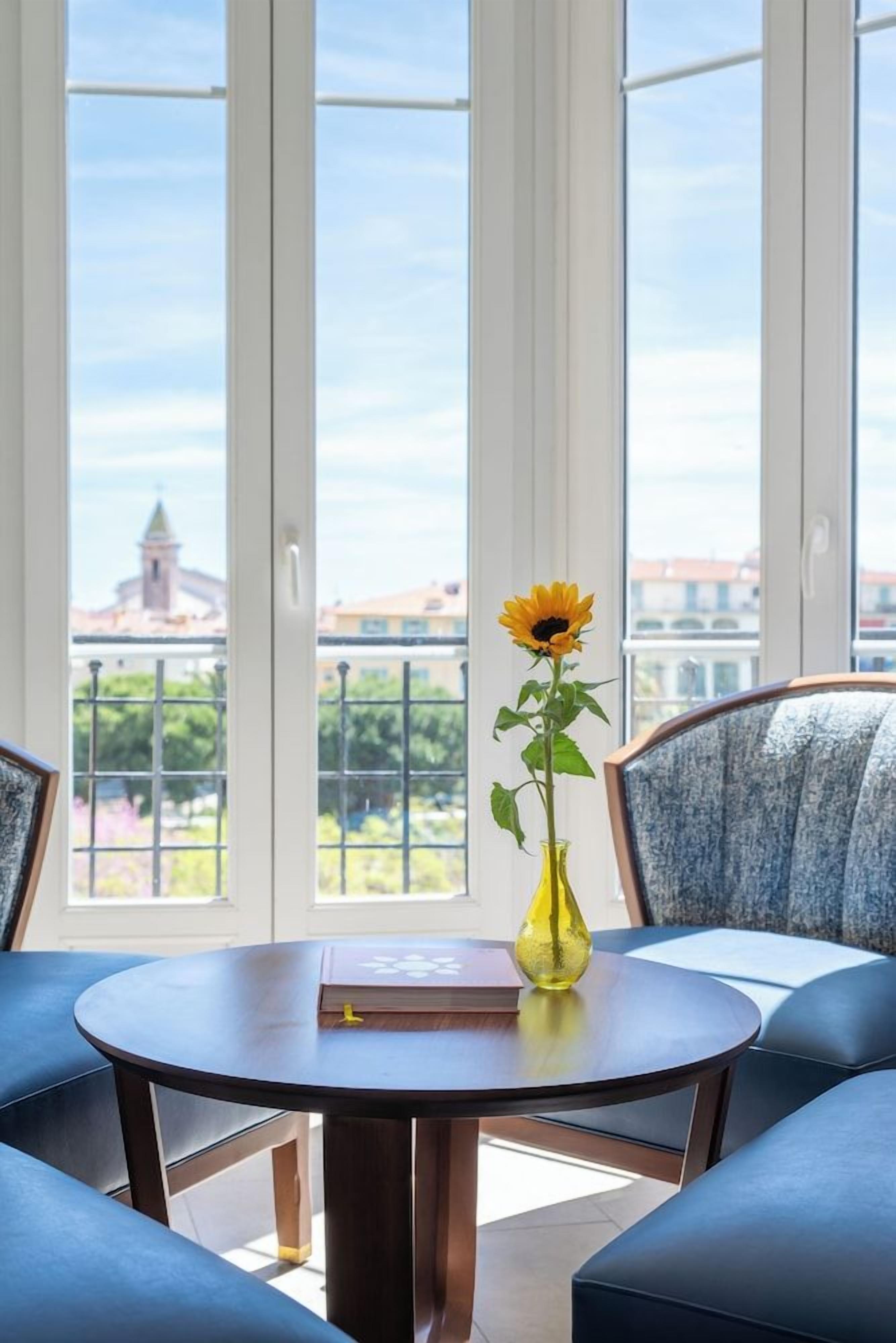 A room with a round table, a vase with a sunflower, and a view of buildings through large windows.