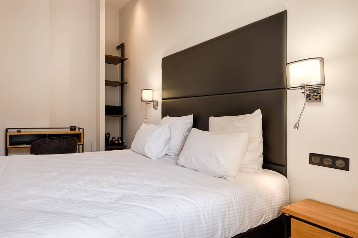 Ein modernes Hotelzimmer mit einem großen Bett, einem Schreibtisch und einem Sessel. An den Wänden sind Lampen angebracht, neben dem Bett steht ein kleiner Tisch.