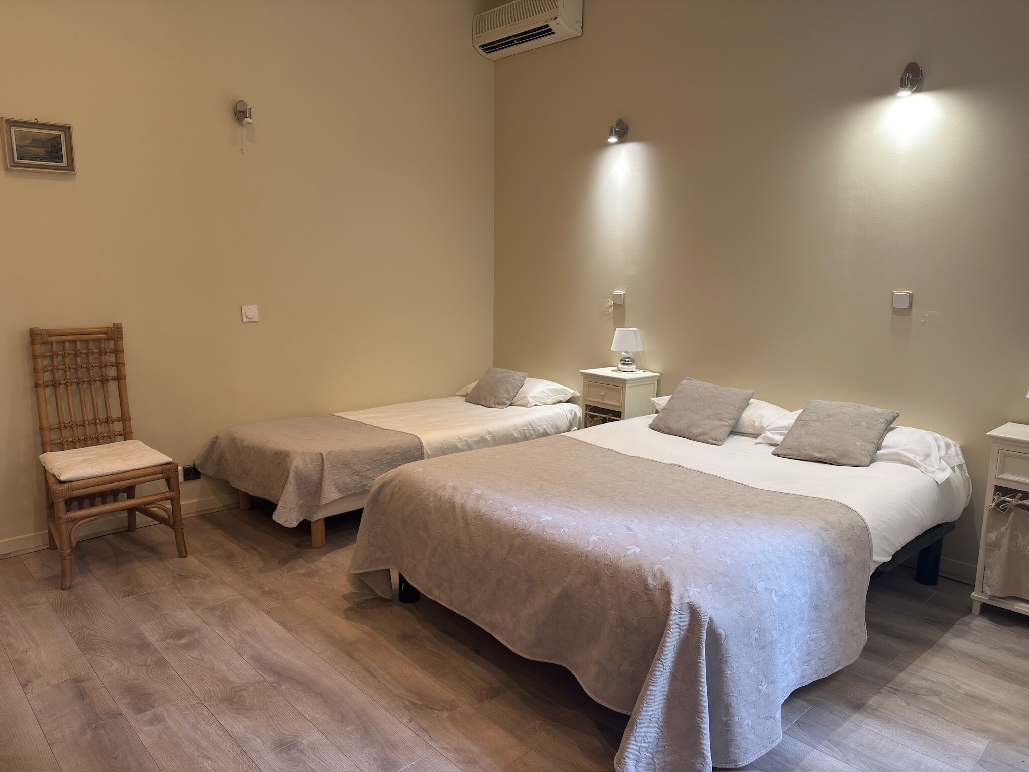 Habitación de hotel con dos camas individuales, una silla de madera, una mesita de noche con lámpara y un aire acondicionado instalado en la pared.