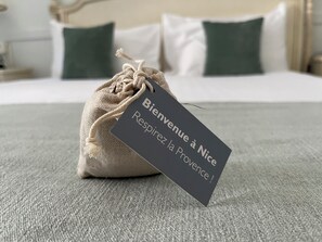 Un petit sac en toile de jute avec une étiquette sur laquelle on peut lire : « Bienvenue à Nice, respirez l’air de la Provence ! », posé sur un lit avec une literie blanche et des coussins verts.