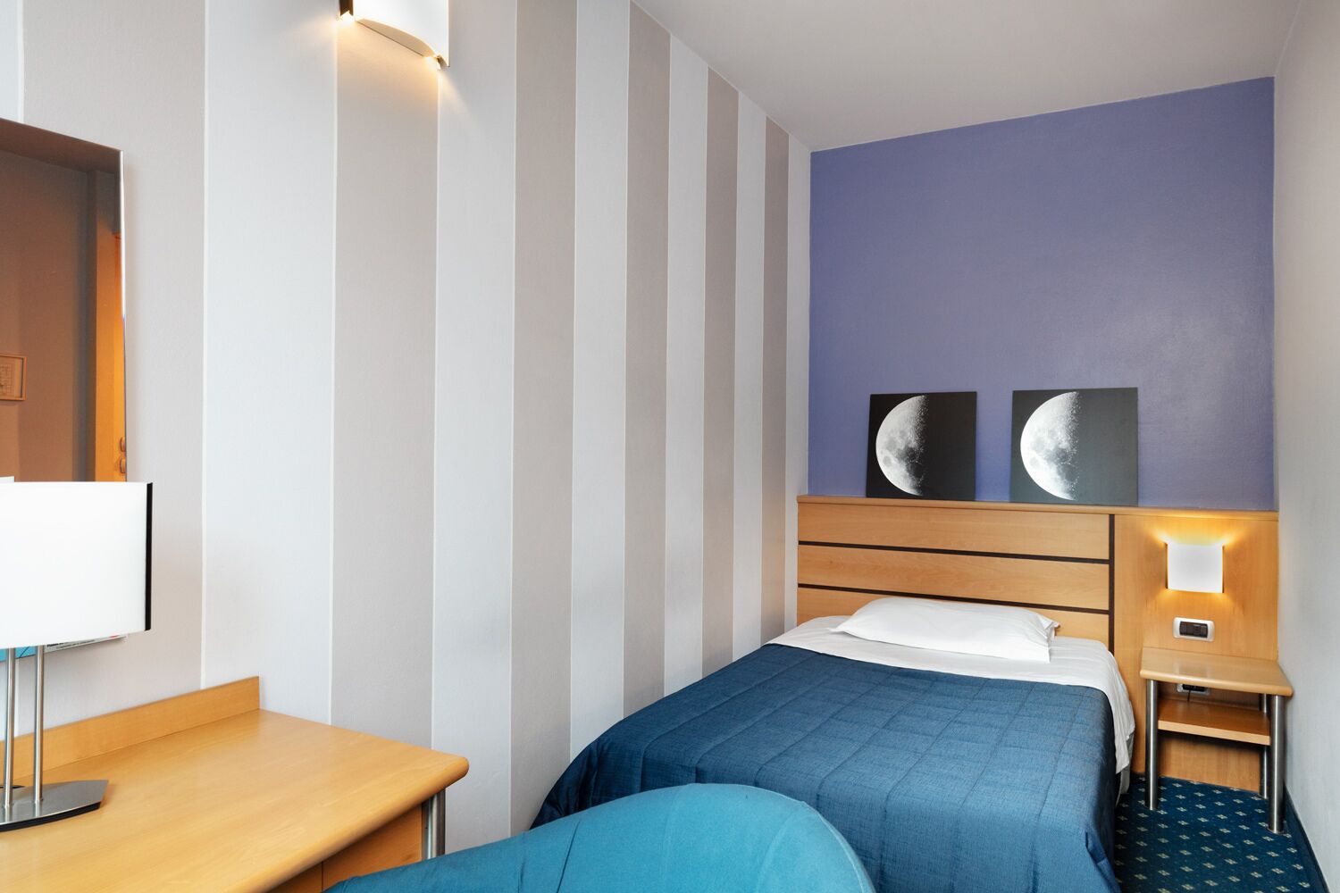 A hotel room with a bed, a desk, and a nightstand. There are two wall-mounted picture frames and a wall-mounted light fixture.