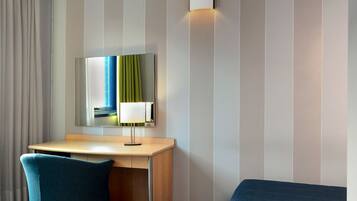 A hotel room with a desk, a chair, a bed, a mirror, and a wall-mounted lamp.