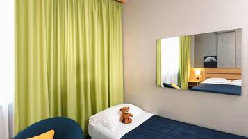 Une chambre d’hôtel avec un lit, une chaise, une petite table sur laquelle est posé un ours en peluche, et un miroir qui reflète le lit, ainsi qu’une table de chevet avec une lampe.
