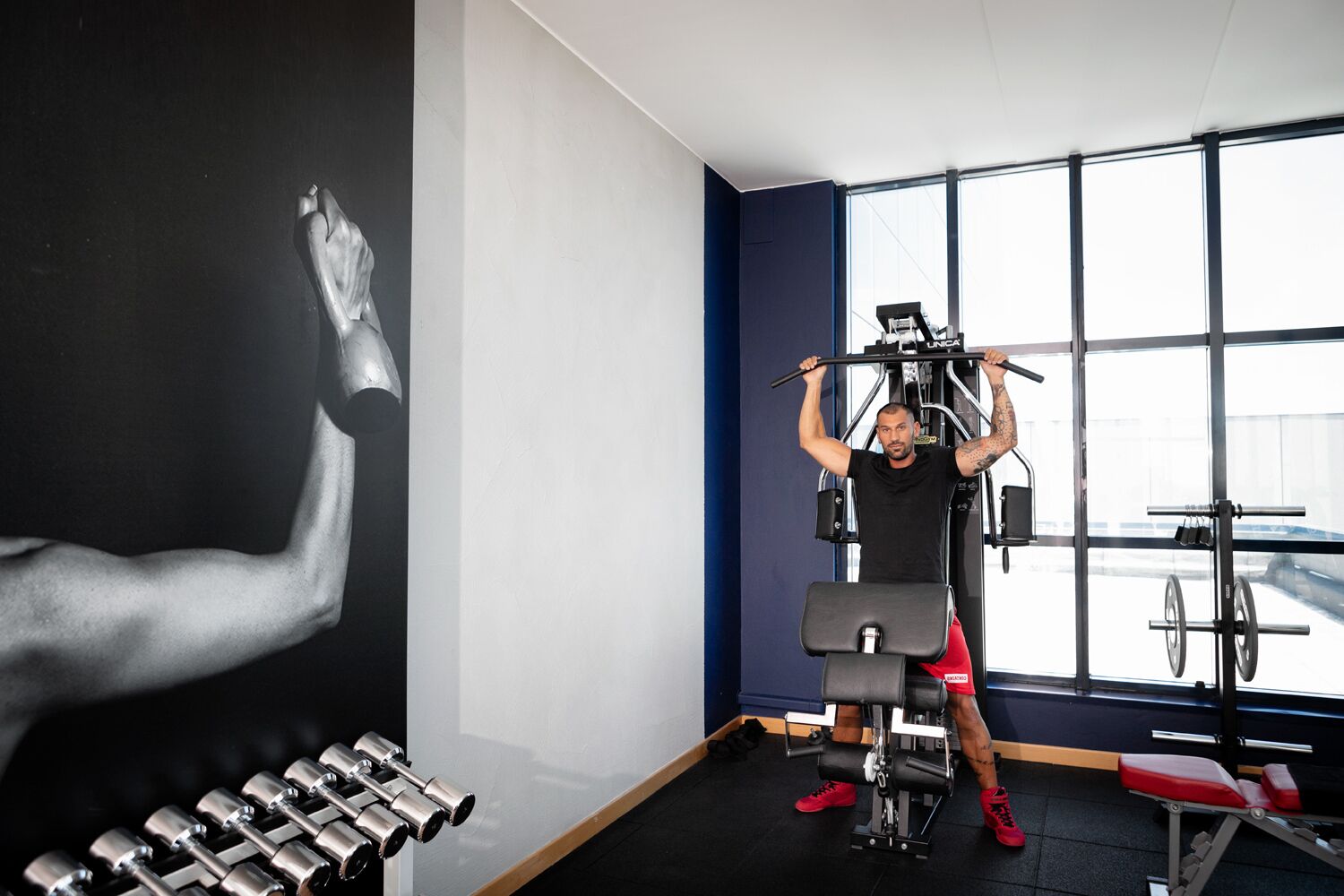 A gym with a person using a cable machine, a wall with a muscular arm illustration, and a window with a view of a car.