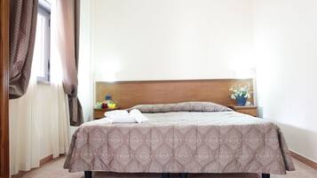 Un letto ben rifatto con una coperta a motivi, una testiera in legno, un comodino con un vaso di fiori e una finestra con tende.