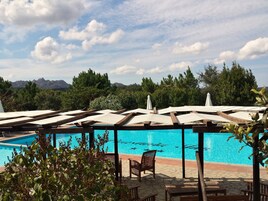 Zona de piscina con sillas de madera y sombrillas, rodeada de vegetación y montañas al fondo.