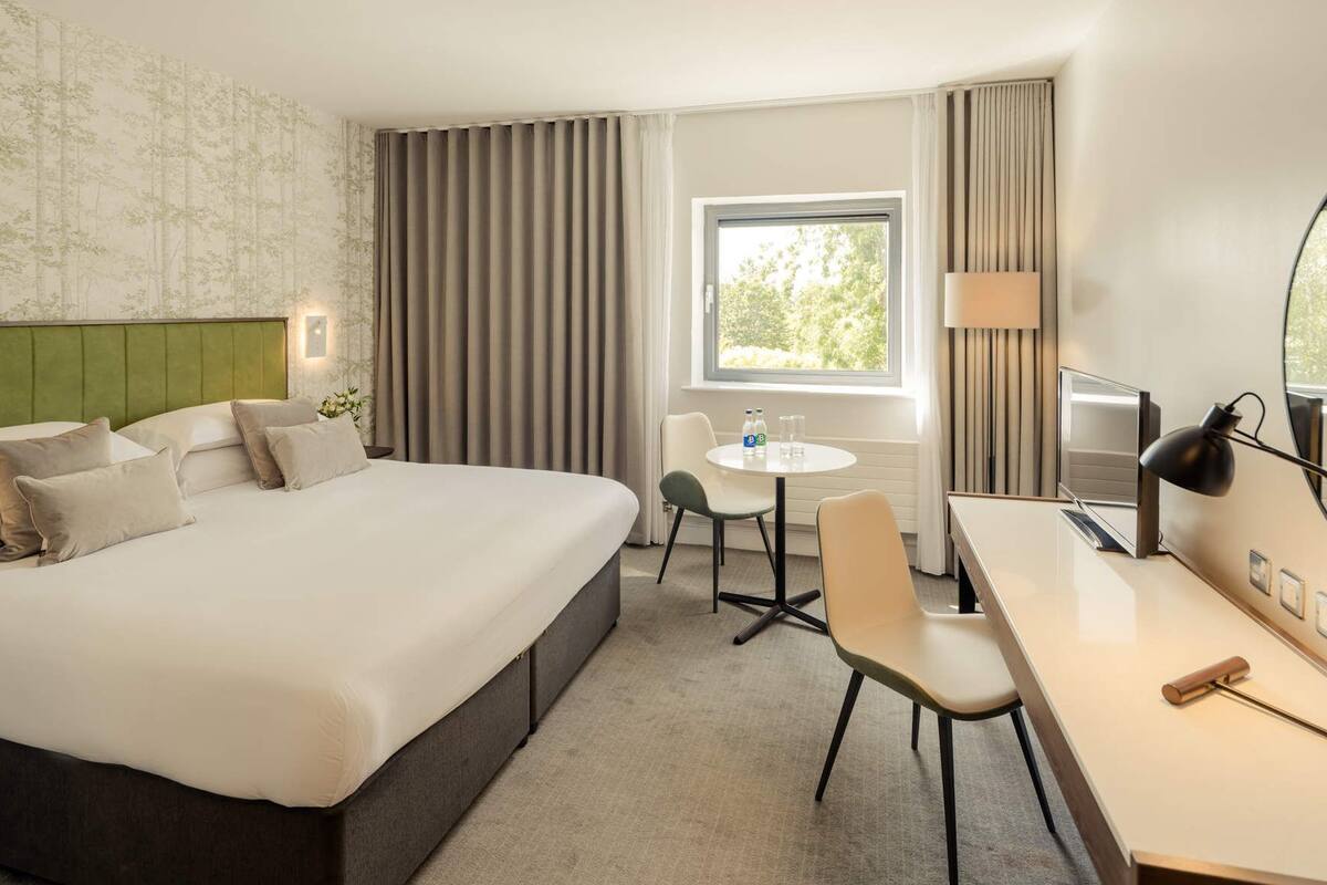 A hotel room with a large bed, a desk, a chair, a small table, a lamp, and a window with curtains.
