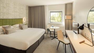 A hotel room with a large bed, a desk, a chair, a small table, a lamp, and a window with curtains.