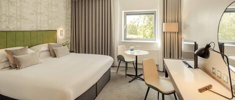 A hotel room with a large bed, a desk, a chair, a small table, a lamp, and a window with curtains.