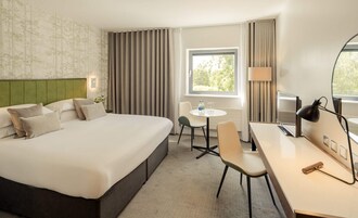 Habitación de hotel con una cama grande, un escritorio, una silla, una mesita, una lámpara y una ventana con cortinas.