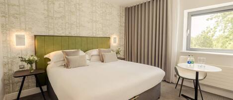 Une chambre d’hôtel avec un grand lit, une table de chevet ornée d’un vase de fleurs, une petite table ronde avec deux bouteilles et des verres, et une fenêtre avec des rideaux.