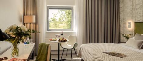 Una habitación de hotel con cama, escritorio con silla, ventana con cortinas y una mesa con un florero y una botella de vino.