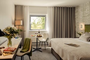 Una habitación de hotel con cama, escritorio con silla, ventana con cortinas y una mesa con un florero y una botella de vino.