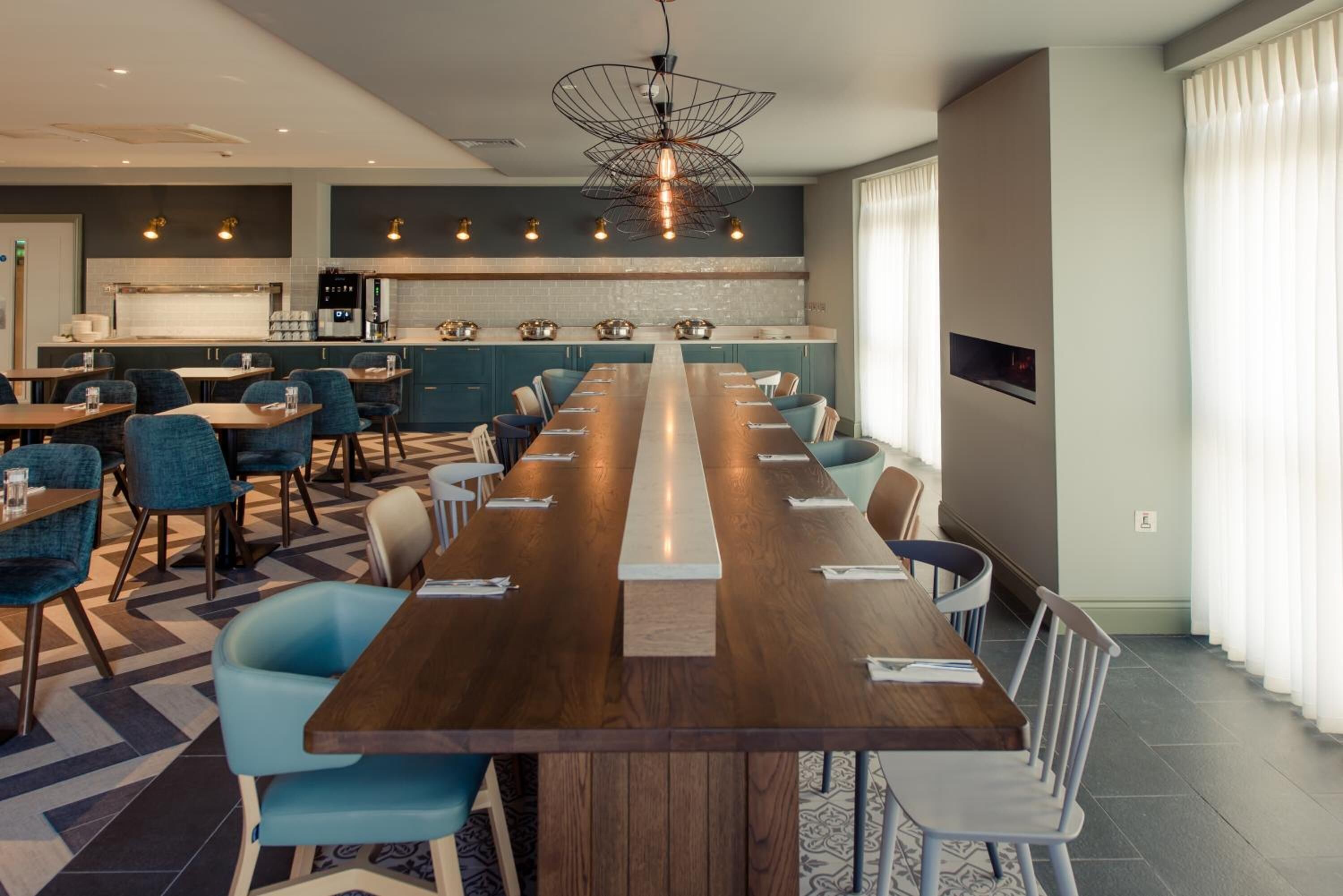 A modern dining area with a long wooden table, blue chairs, and a kitchen in the background.