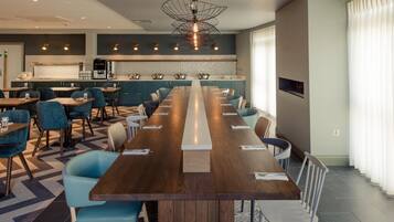 A modern dining area with a long wooden table, blue chairs, and a kitchen in the background.