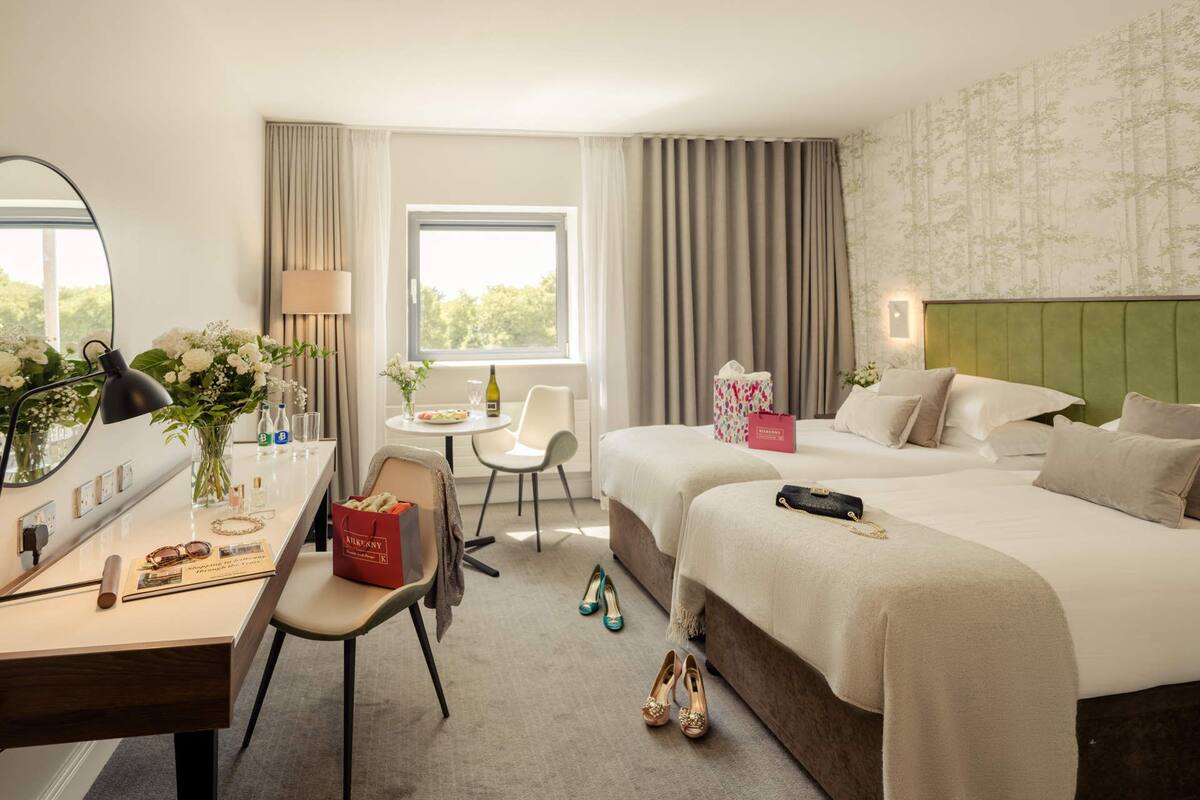 A hotel room with a bed, a desk, a chair, a table with a vase of flowers, and a window with curtains.