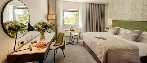 A hotel room with a bed, a desk with a chair, a mirror, a window with curtains, and a wall with a patterned wallpaper.