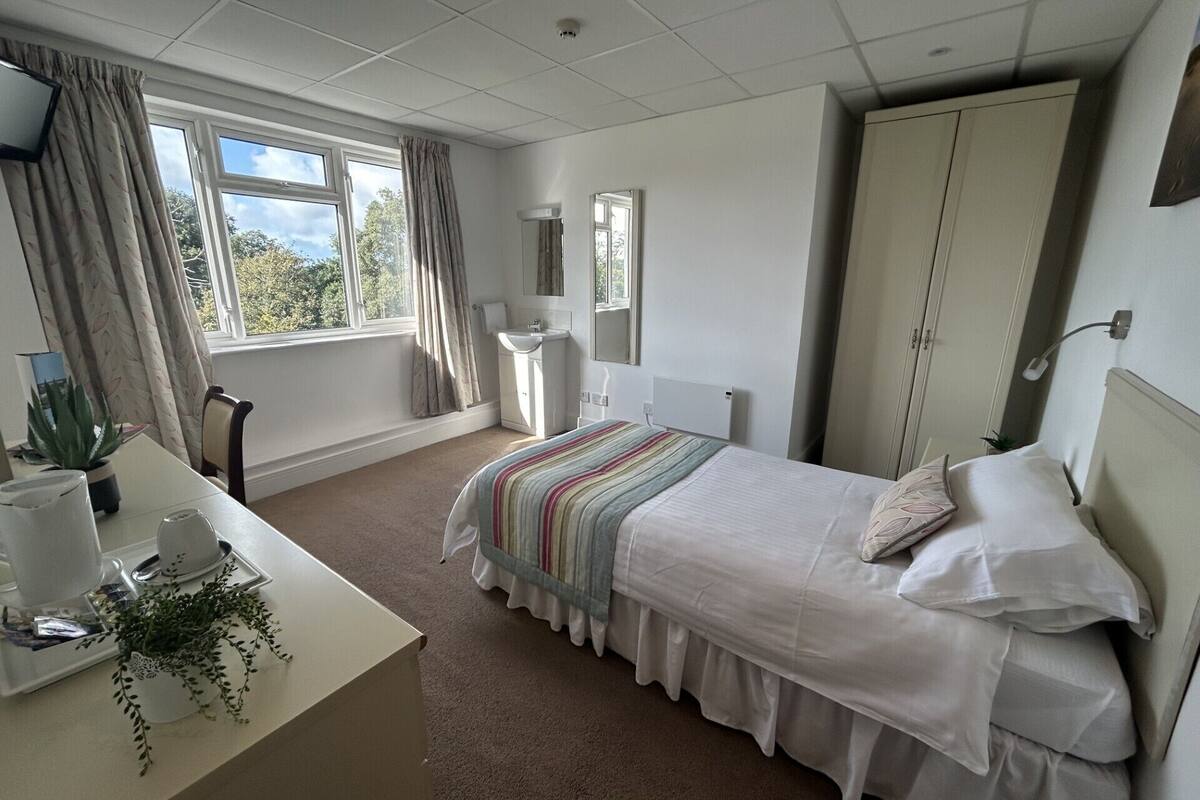 A bedroom with a bed, a desk, a chair, a wardrobe, a window with curtains, and a potted plant.