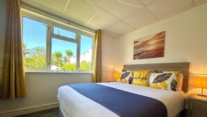 Hotelkamer met een bed, een raam met gordijnen, een schilderij aan de muur en een nachtkastje met een lamp.