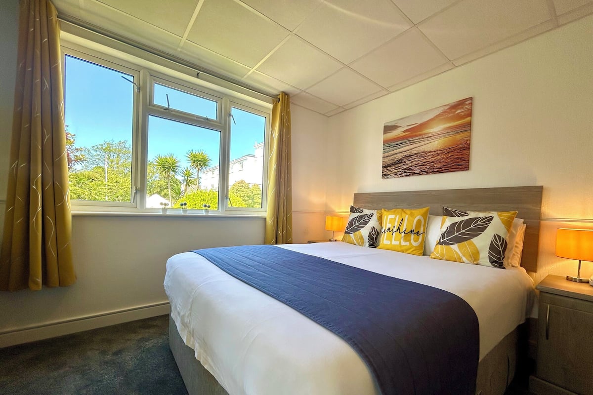 A hotel room with a bed, a window with curtains, a painting on the wall, and a nightstand with a lamp.