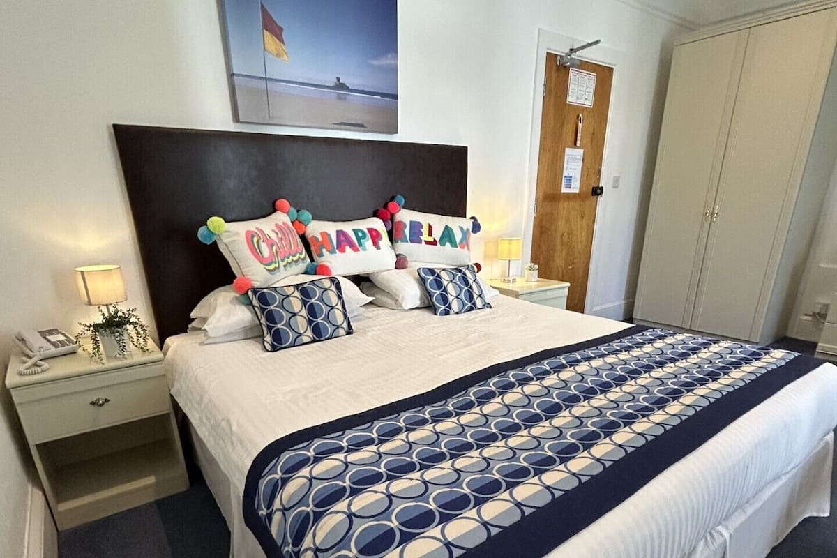 A neatly made bed with a patterned bedspread, a headboard, and decorative pillows with text. There is a bedside table with a lamp, a small plant, and a phone. A door with a sign is visible in the background.