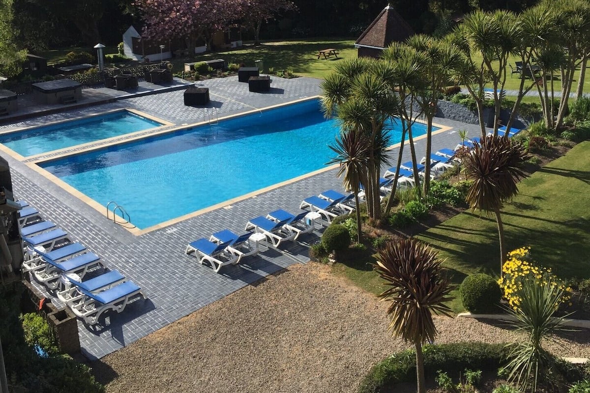 A rectangular swimming pool with a tiled deck, surrounded by lounge chairs and palm trees.