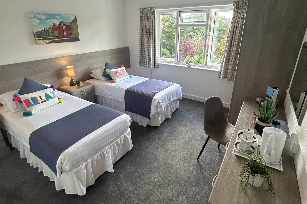 A hotel room with two beds, a desk, a chair, a window with curtains, a painting on the wall, and a small plant on the desk.